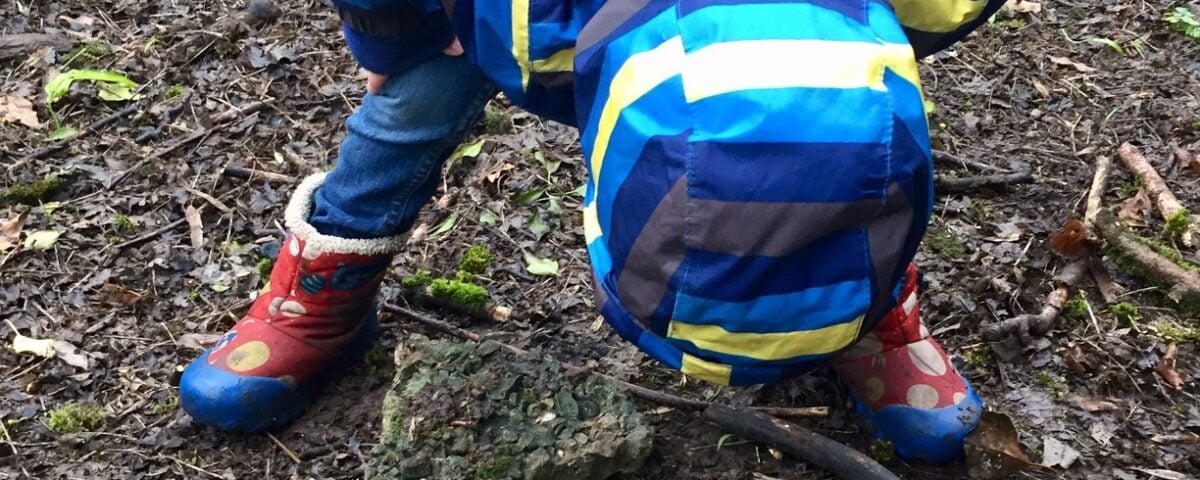 A child wearing blue coat and rainbow wellies, bent over examining the ground