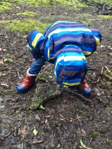 A child wearing blue coat and rainbow wellies, bent over examining the ground