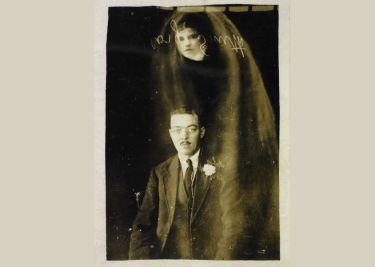 A man in a suit sits in front of a dark background, while a ghostly, transparent figure of a woman with a veil appears behind him.