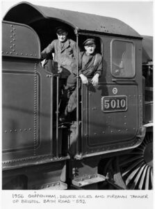 A black-and-white photo shows two men in work clothes and caps on a train. One man stands on the steps of the train's cab, while the other leans out of a window. The train has a number plate with "5010" on its side.