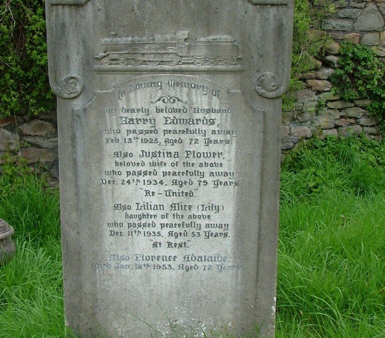 An ornate, arched grey stone headstone stands upright in a lush green grassy field in front of a rustic stone wall. At the top of the headstone, a detailed carving of a vintage steam locomotive train sits above the primary inscription text. The memorial inscription is dedicated to Harry Edwards (died 1925), his wife Justina Flower (died 1934), their daughter Lilian Alice (Lily) (died 1935), and Florence Adelaide (died 1953). Green ivy and leafy bushes frame the top and sides of the stone wall behind the grave marker.