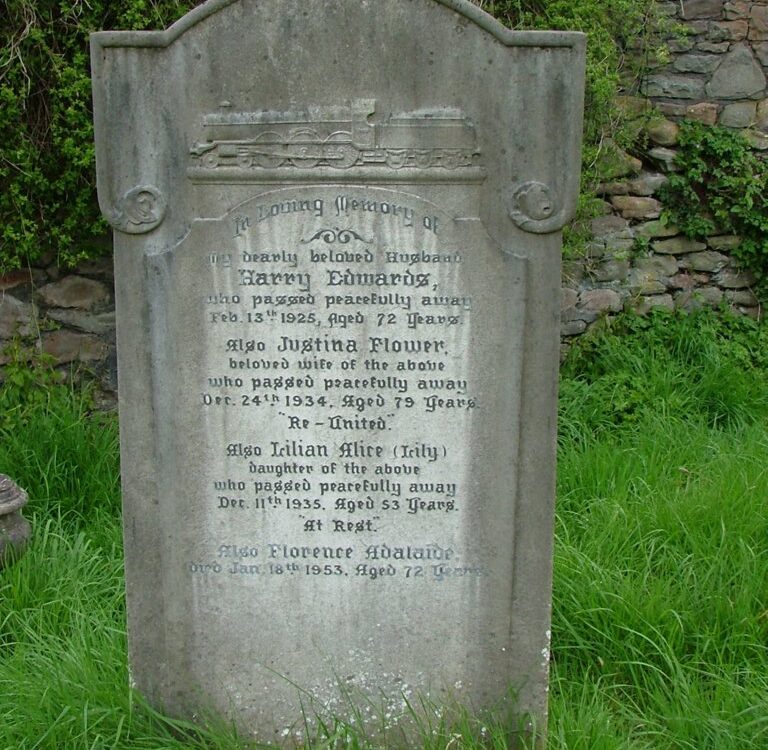 An ornate, arched grey stone headstone stands upright in a lush green grassy field in front of a rustic stone wall. At the top of the headstone, a detailed carving of a vintage steam locomotive train sits above the primary inscription text. The memorial inscription is dedicated to Harry Edwards (died 1925), his wife Justina Flower (died 1934), their daughter Lilian Alice (Lily) (died 1935), and Florence Adelaide (died 1953). Green ivy and leafy bushes frame the top and sides of the stone wall behind the grave marker.