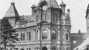 A black-and-white photograph of a large, ornate building with a central clock tower topped by a weather vane. The building features arched entrances, multiple turrets, and decorative stonework, standing next to a paved street.